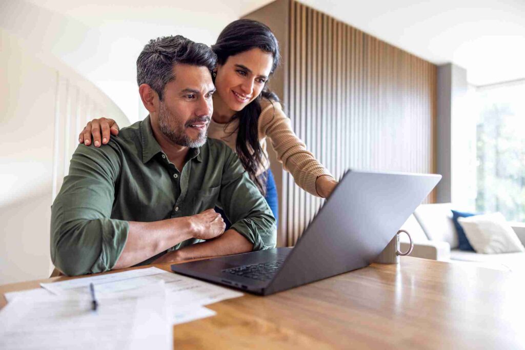 A couple smiling while looking at a laptop screen together at a table.