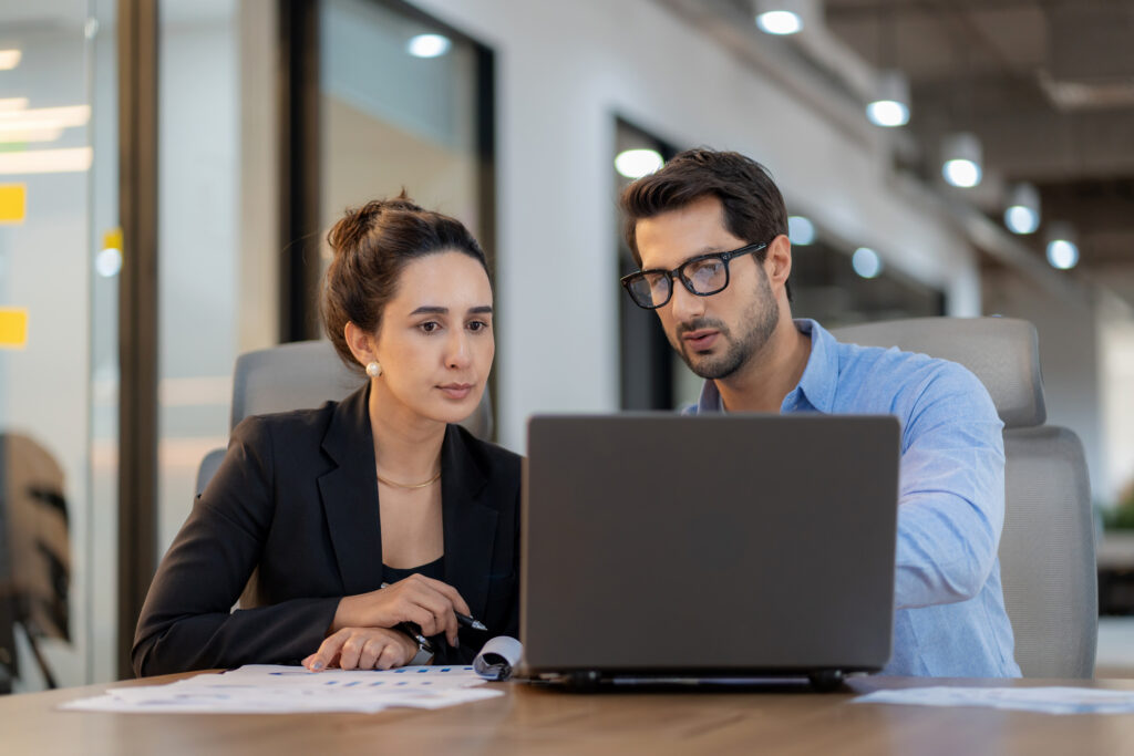 A woman in a dark suit sitting at a desk with a male colleague looking at a laptop.