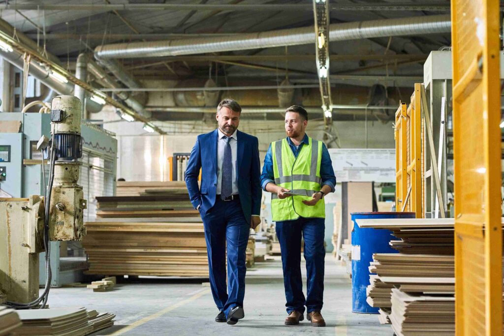 A financial advisor in a blue suit walking through a manufacturing facility alongside a small business owner in a high-visibility safety vest and hard hat.